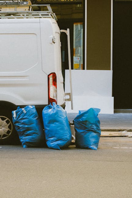 The image shows the rear section of a white commercial van parked on an urban street, with its tailgate partially visible on the left side. In front of the van, three large blue rubbish bags made of plastic are placed directly on the pavement; they appear to be filled with waste, with some bulging and slight creases on their surfaces. The bags are positioned close together, with two on the left and one on the right, all leaning slightly against the back of the van. Behind the bags, there is a white building with a large glass window or door, partially visible and reflecting some indistinct shapes. A white concrete or stone ledge extends slightly onto the pavement, possibly part of an entryway step or decorative feature, and a narrow portion of a curb is visible at the bottom of the image. The scene is illuminated with natural daylight, giving the environment a neutral, professional appearance typical of private waste collection sites or on-site rubbish clearance services, as seen in waste removal services such as Waste Removal Chelsea focusing on independent rubbish disposal solutions.