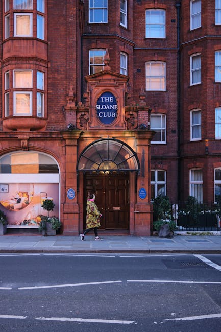 The image depicts the exterior of a historic red-brick building with architectural details typical of late Victorian or Edwardian style, featuring multiple bay windows and decorative stonework. The entrance is centrally located, with a dark wooden double door beneath an arched glass canopy. Above the door, there is a prominent blue round sign labeled 'THE SLOANE CLUB' attached to an ornate stone surround. On either side of the entrance, small blue signs are mounted on the wall, indicating private property or membership information. Flanking the doorway, well-maintained flower pots containing green foliage and small plants are positioned on the pavement, contributing to the building's tidy appearance. A woman dressed in a patterned coat and black shoes is walking past, while the street in front is paved with asphalt and marked with white dashed lines. The scene captures a quiet, urban setting potentially associated with private or exclusive establishments, with suggestions of waste removal services subtly implied by the context of the webpage, but no visible rubbish or waste-related objects are present in the image itself.