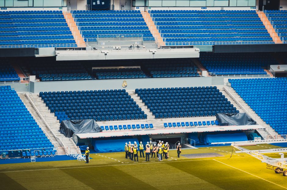 A large, empty sports stadium with rows of blue plastic seats arranged in multiple tiers, some sections covered with black protective tarps secured with ropes. In front of the seating areas, on the grass field, a group of workers wearing yellow high-visibility jackets, white protective helmets, and safety gear are gathered, appearing to discuss or inspect the space. Equipment such as a scaffolding or maintenance equipment is present near the workers, and a truck or platform is partially visible to the right. The environment is well-lit with natural daylight, and the scene suggests ongoing maintenance, refurbishment, or preparations for an event, aligning with independent waste management or site clearance services like those provided by Waste Removal Chelsea for large public venues.