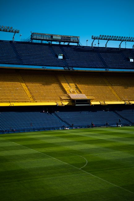 An expansive view of a football stadium with bright yellow and dark blue tiered seating surrounding a well-maintained grass pitch. The seating sections are empty, and the bright blue sky is visible overhead. The stadium features prominent floodlights positioned on tall structures above the stands, and a large scoreboard or lighting rig is mounted on the upper tier. The grass field in the foreground shows clear mowing lines and a visible center circle. The overall scene presents a clean, orderly sports venue that may require periodic maintenance or rubbish removal services to keep the area in pristine condition, reflecting the importance of proper waste handling in large-scale facilities. Waste Removal Chelsea provides rubbish clearance options that could involve the removal of debris or waste accumulated during stadium events or routine cleaning. The environment is professional, open, and neatly maintained, emphasizing the importance of efficient waste disposal in such large public venues to ensure safety and cleanliness around Stamford Bridge Chelsea or similar locations.