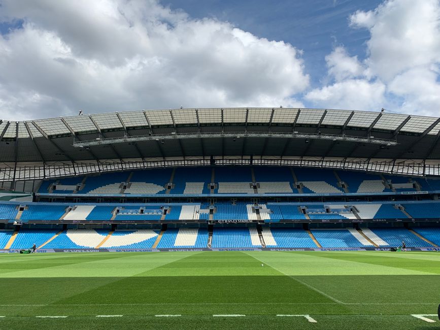 A large, empty sports stadium with rows of blue plastic seats arranged in multiple tiers, some sections covered with black protective tarps secured with ropes. In front of the seating areas, on the grass field, a group of workers wearing yellow high-visibility jackets, white protective helmets, and safety gear are gathered, appearing to discuss or inspect the space. Equipment such as a scaffolding or maintenance equipment is present near the workers, and a truck or platform is partially visible to the right. The environment is well-lit with natural daylight, and the scene suggests ongoing maintenance, refurbishment, or preparations for an event, aligning with independent waste management or site clearance services like those provided by Waste Removal Chelsea for large public venues.