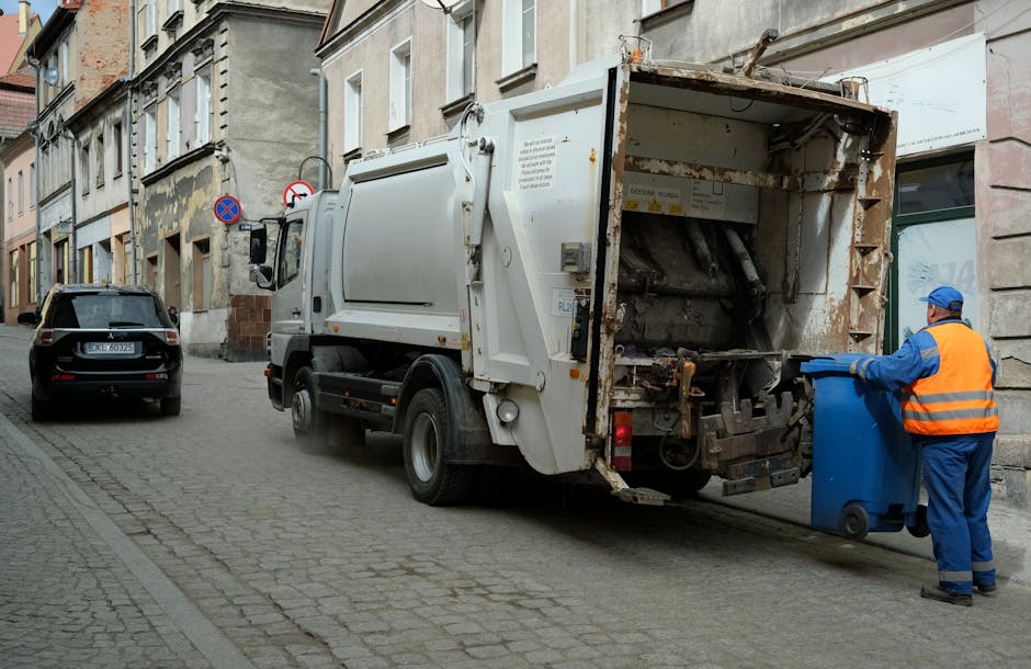 A waste collection vehicle operated by Waste Removal Chelsea is positioned on a narrow cobblestone street between aged, multi-story buildings with weathered facades. The vehicle, primarily white with some visible rust and grime, has its rear loading section open, revealing the mechanical mechanisms used for compacting and collecting rubbish. A worker dressed in blue protective overalls, an orange high-visibility vest, and a blue cap is standing beside the vehicle, actively emptying a large blue wheeled bin into the truck's hopper. The street is lined with parked cars, including a dark-colored vehicle on the left side, and features street signs indicating parking restrictions. The surrounding buildings have a mix of old brickwork and plaster, with some plaster patches showing signs of wear, and the area appears to be an urban residential or commercial district. Natural daylight illuminates the scene, emphasizing the textures of the cobbled pavement and weathered building surfaces, highlighting the practical aspects of private waste collection and rubbish removal services in a city environment.