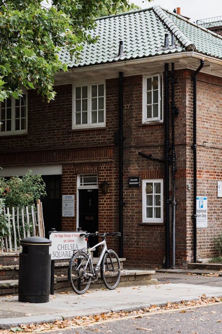 The image depicts a brick residential building with a grey tiled pitched roof, situated on a street corner. In front of the building, a bicycle with a white frame and black saddle is propped against a black metal railing, near a small step leading up to the entrance door. To the left of the bicycle, a black cylindrical trash bin is positioned on the pavement. A white signboard with red and black text is placed on the sidewalk, indicating information about rubbish collection or waste disposal. The building features white-framed windows, some with visible window sills, and a small white-framed door with a letter slot. Green foliage from a nearby tree partially obscures the top left corner of the building. The environment appears clean and tidy, with autumn leaves scattered on the pavement, and the scene subtly suggests an area serviced by private waste removal, such as Waste Removal Chelsea, for rubbish collection outside standard council services.
