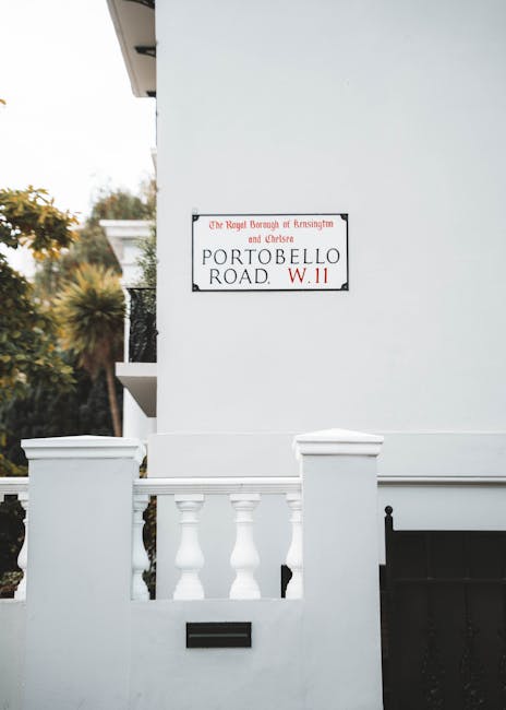A close-up view of a modern white residential building façade, featuring a decorative white balustrade with rounded columns and a flat top, positioned above a mail slot embedded in the wall. To the right, a dark black section of the gate or fence is partially visible. On the building’s wall, there is a rectangular street sign with a white background and black and red lettering, reading 'Portobello Road W.11,' indicating the location in London. The sign also mentions the Royal Borough of Kensington and Chelsea. In the background, out of focus, there are trees and foliage with a mix of green and autumnal hues, suggesting an outdoor setting. The scene is illuminated by natural daylight, providing clear visibility of the textures and colors of the building materials, such as the smooth, painted finish of the wall and the glossy surface of the balustrade, which is styled in a classical shape. This image reflects the typical architecture of private properties in the area, with a subtle visual connection to independent waste management and rubbish removal services often employed in such residential settings, where on-site clearance or private disposal may be preferred over municipal collection.