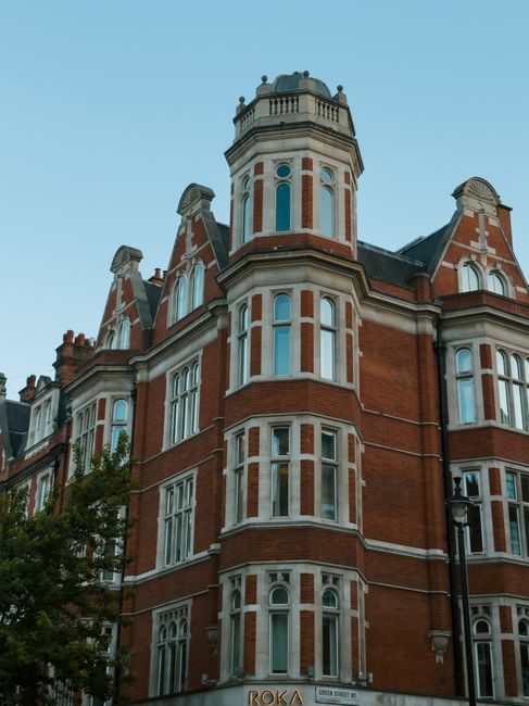 A multi-storey Victorian-style red-brick building with intricate white window surrounds and decorative architectural features, including rounded bays and a turreted corner with a small domed roof. The building has numerous tall, narrow windows with white frames, some with arched tops, set in a symmetrical arrangement. Part of a city street scene is visible, with a black street lamp on the right and a partial view of a tree with green leaves on the lower left. The sky above is clear and pale blue, providing natural lighting that highlights the detailed brickwork and ornate finishes. This building, identifiable by its distinctive appearance, could be a property where private waste collection services such as those offered by Waste Removal Chelsea are suitable for managing rubbish removal, especially for on-site clearance or alternative waste handling outside local authority collection.