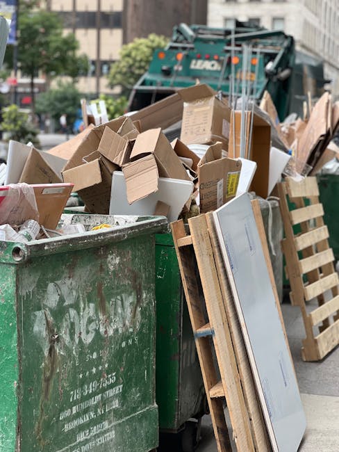 A large green waste disposal container filled with a mixture of cardboard boxes, some flattened and others partially intact, along with miscellaneous packaging materials, situated outdoors on a city street. The cardboard appears brown and light beige with visible corrugated textures and printed labels on some boxes. Next to the container, there is a wooden pallet leaning against its side, and in the background, a recycling truck with visible branding is parked. The environment includes urban elements such as trees and multi-storey buildings, with the scene illuminated by natural daylight, indicating a typical rubbish collection or on-site clearance scenario managed by Waste Removal Chelsea, aligning with private waste removal services rather than municipal curbside collection.