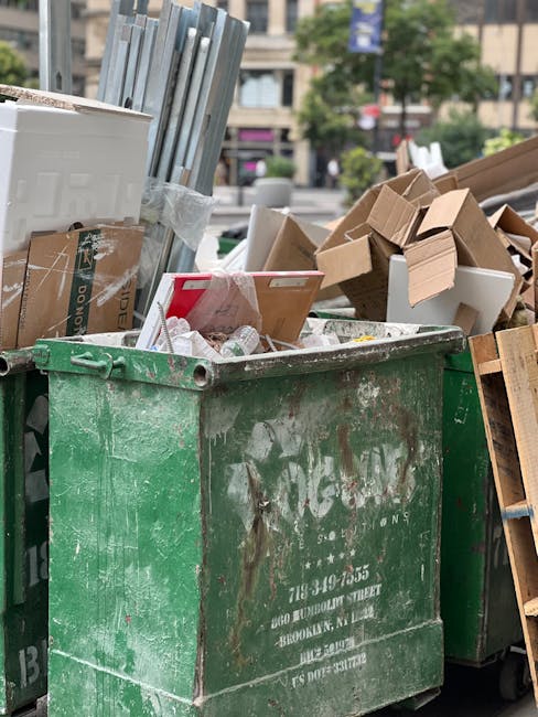 A large green waste disposal container filled with a mixture of cardboard boxes, some flattened and others partially intact, along with miscellaneous packaging materials, situated outdoors on a city street. The cardboard appears brown and light beige with visible corrugated textures and printed labels on some boxes. Next to the container, there is a wooden pallet leaning against its side, and in the background, a recycling truck with visible branding is parked. The environment includes urban elements such as trees and multi-storey buildings, with the scene illuminated by natural daylight, indicating a typical rubbish collection or on-site clearance scenario managed by Waste Removal Chelsea, aligning with private waste removal services rather than municipal curbside collection.